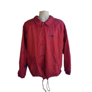 May include: A red coach jacket with a button-up front, long sleeves, and a drawstring hem. The jacket has a small black patch on the chest and a blue patch on the left sleeve. The jacket is displayed on a mannequin.