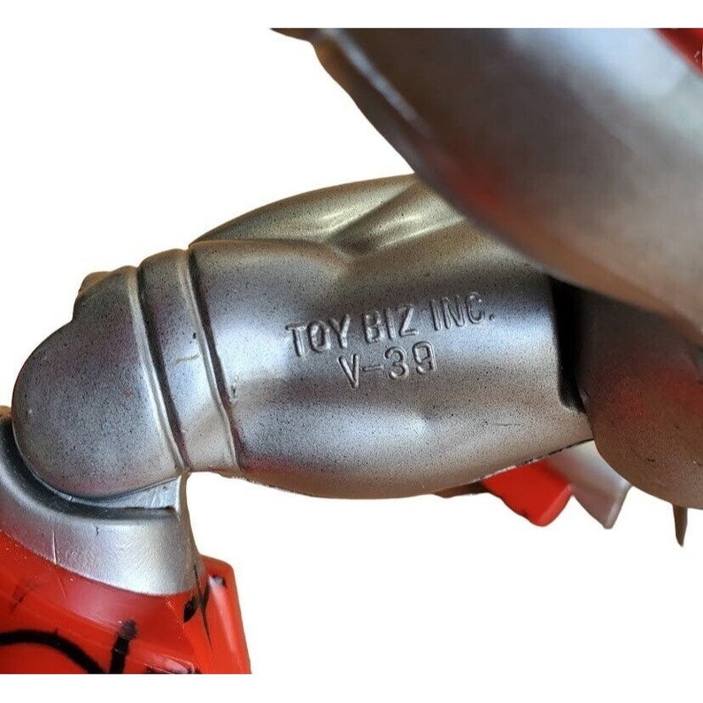 K&ouml;nnte beinhalten: Nahaufnahme eines silberfarbenen Plastikspielzeugarms mit der Aufschrift "TOY BIZ INC. V-39".