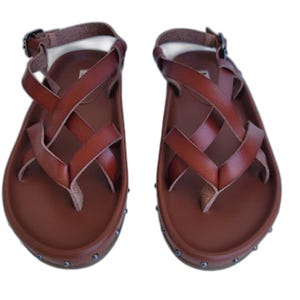 May include: A pair of brown leather sandals featuring a thong-style toe post, crisscrossing straps across the top, and an adjustable ankle strap with a metal buckle. The footbed is contoured, and the base is accented with small metal studs.