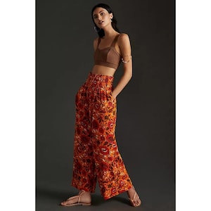 K&ouml;nnte beinhalten: Ein Damen-Outfit mit einem braunen Crop-Top und einer weiten Hose. Die Hose ist leuchtend orange mit einem Blumenmuster in Rot und Gelb. Das Outfit wird mit Sandalen kombiniert.