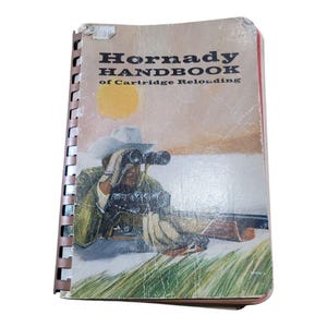 May include: Vintage "Hornady Handbook of Cartridge Reloading" book. The cover features a cowboy with binoculars, a rifle, and a sunset background. The book has a spiral binding and shows signs of wear.