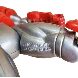 K&ouml;nnte beinhalten: Nahaufnahme einer silbernen und roten Actionfigur mit dem Text "&copy; 1995 MARVEL MADE IN CHINA" auf dem Arm der Figur.