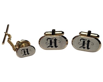 May include: A set of gold-toned cufflinks and a tie tack. Each piece features a silver oval with a black letter 'H' in a decorative font.