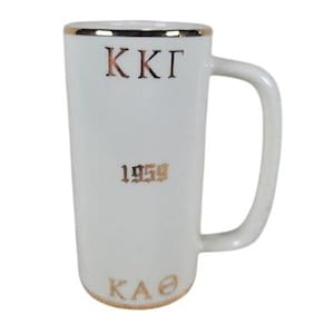 May include: White ceramic mug with a gold rim and handle. The mug features the letters "KKΓ", the number "1959", and the letters "KAΘ" in gold lettering. The mug is tall and cylindrical.