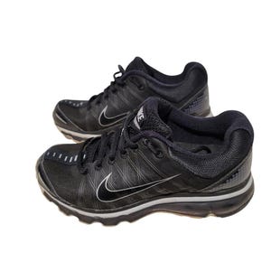 May include: Pair of black Nike athletic shoes. The shoes feature a black upper with a white Nike swoosh logo on the sides. The soles are black and gray. The shoes have black laces and a padded collar.