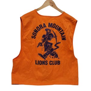 May include: Orange sleeveless vest with a blue graphic of a cartoon cat wearing a hat and holding a pickaxe. The graphic is surrounded by a blue circle with the text "Sonora Mountain Lions Club".