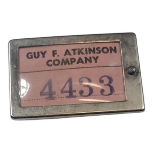 May include: A rectangular metal tag with the text "GUY F. ATKINSON COMPANY" above the number "4433". The background is a light pink color, and the numbers are a dark purple. The tag has a silver metal frame.