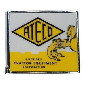 May include: A rectangular pin with a white background and a yellow and black diamond shape with the word "ATECO". Below the diamond is a yellow bar with the text "AMERICAN TRACTOR EQUIPMENT CORPORATION". A yellow tractor arm is on the right.