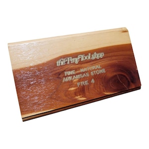 May include: A rectangular sharpening stone from "The Fine Tool Shop." The top is a mix of light and dark wood, with the text "FINE - NATURAL ARKANSAS STONE FRE 4" etched into the darker wood. The wood grain is visible.