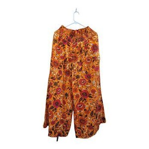 K&ouml;nnte beinhalten: Weithose mit orangefarbenem Blumenmuster, h&auml;ngend an einem wei&szlig;en Kleiderb&uuml;gel. Die Hose zeigt ein sich wiederholendes Design aus roten und rosa Blumen mit gr&uuml;nen Bl&auml;ttern auf orangefarbenem Hintergrund. Der Bund ist gerafft, und der Stoff wirkt leicht.