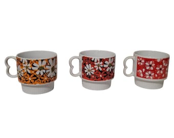 May include: Three white ceramic mugs with heart-shaped handles. Each mug has a different floral pattern: orange and black with white flowers, red and black with white flowers, and red with white flowers.