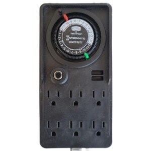 May include: A black rectangular electrical power strip with a built-in analog timer dial at the top. The dial features the text Intermatic Heavy Duty and Time of Day. It includes a red pin, a green pin, a circular button, and six electrical outlets.