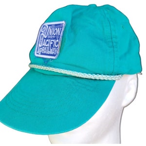 May include: A turquoise blue baseball cap with a white rope trim and a blue and white patch that says "Union Pacific Railroad".