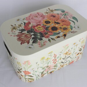 May include: Cream-colored rectangular storage box with a floral design. The lid features a bouquet of pink, orange, and yellow flowers. The sides are decorated with wildflowers. Includes a black handle.