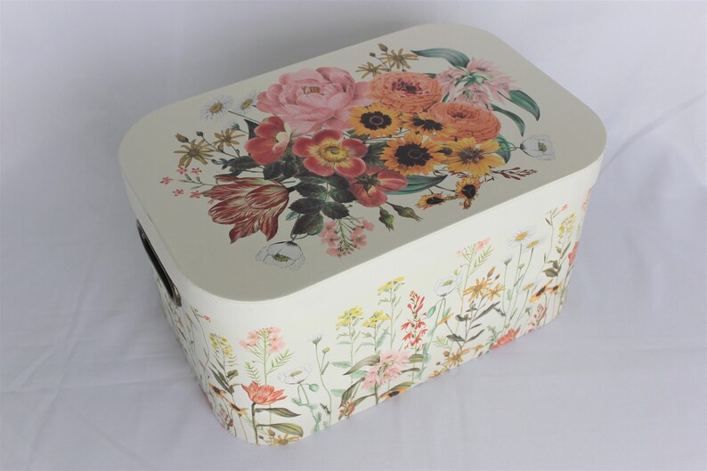 May include: Cream-colored rectangular storage box with a floral design. The lid features a bouquet of pink, orange, and yellow flowers. The sides are decorated with wildflowers. Includes a black handle.