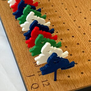 May include: Wooden board game with a series of holes and colorful peg pieces. The pegs are shaped like skiers in red, white, green, and blue. The board has the numbers 6, 9, 10, 11, and 12 etched into the wood.