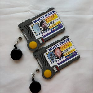 Puede incluir: Dos tarjetas multi-pass de pl&aacute;stico gris con una foto en blanco y negro de una persona en cada tarjeta. Las tarjetas tienen un bot&oacute;n amarillo y un cord&oacute;n retr&aacute;ctil negro.