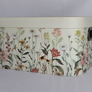 May include: Rectangular storage box with a cream-colored lid and floral pattern. The box features a variety of wildflowers in shades of pink, yellow, and white against a light background. It has a black handle on the side.