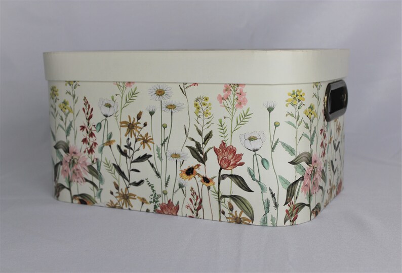 May include: Rectangular storage box with a cream-colored lid and floral pattern. The box features a variety of wildflowers in shades of pink, yellow, and white against a light background. It has a black handle on the side.