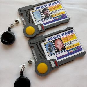 Puede incluir: Dos tarjetas multi-pass de pl&aacute;stico gris con fotos en blanco y negro de personas y texto que dice "MULTI PASS" y "KORBEN DALLAS" y "LEELOO DALLAS". Las tarjetas est&aacute;n unidas a carretes de identificaci&oacute;n retr&aacute;ctiles negros.