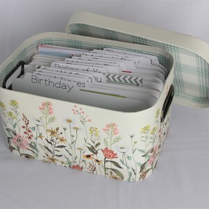 May include: Cream-colored card box with a floral design featuring pink, yellow, and white flowers. The box has a matching lid with a green and white plaid interior. The box contains cards with labeled tabs, including "Birthday" and "Thinking of You."