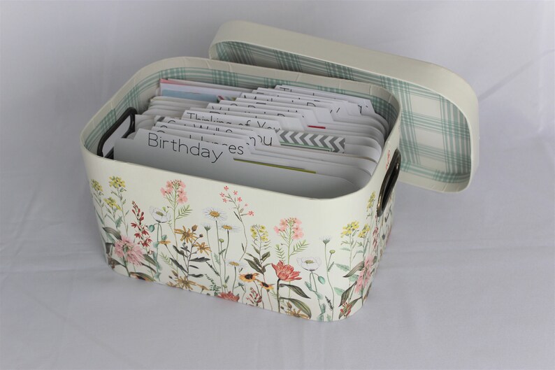 May include: Cream-colored card box with a floral design featuring pink, yellow, and white flowers. The box has a matching lid with a green and white plaid interior. The box contains cards with labeled tabs, including "Birthday" and "Thinking of You."