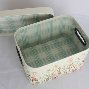 May include: Cream-colored rectangular storage basket with a matching lid. The interior features a light green and white plaid pattern. The exterior is decorated with a floral design. The basket has black handles.