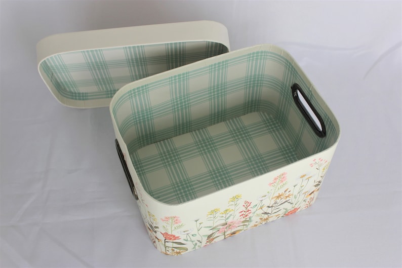 May include: Cream-colored rectangular storage basket with a matching lid. The interior features a light green and white plaid pattern. The exterior is decorated with a floral design. The basket has black handles.