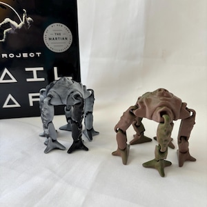 May include: Two articulated, insect-like figurines in gray and brown hues stand before a book. The gray figurine has black accents, while the brown one features green and pink details. The book's cover is black with white text.