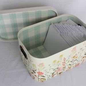 May include: A rectangular storage box with a lid, decorated with a floral pattern. The interior and lid lining feature a green and white plaid design. The box contains labeled cards, and has black handles.