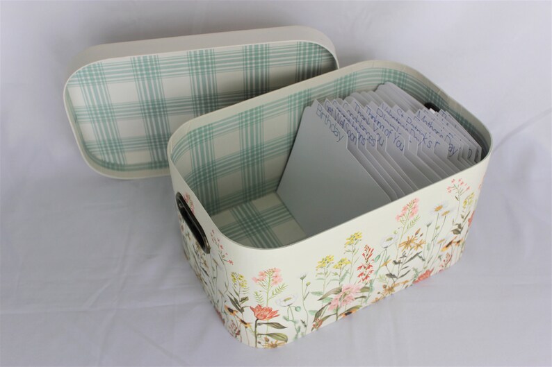 May include: A rectangular storage box with a lid, decorated with a floral pattern. The interior and lid lining feature a green and white plaid design. The box contains labeled cards, and has black handles.
