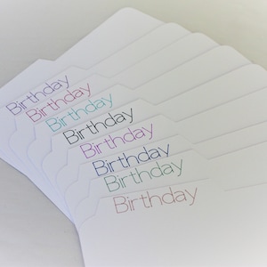 May include: A stack of white file folder tabs with the word "Birthday" printed in various colors. The tabs are arranged in a fan shape, with the text in purple, pink, teal, black, blue, green, and red.