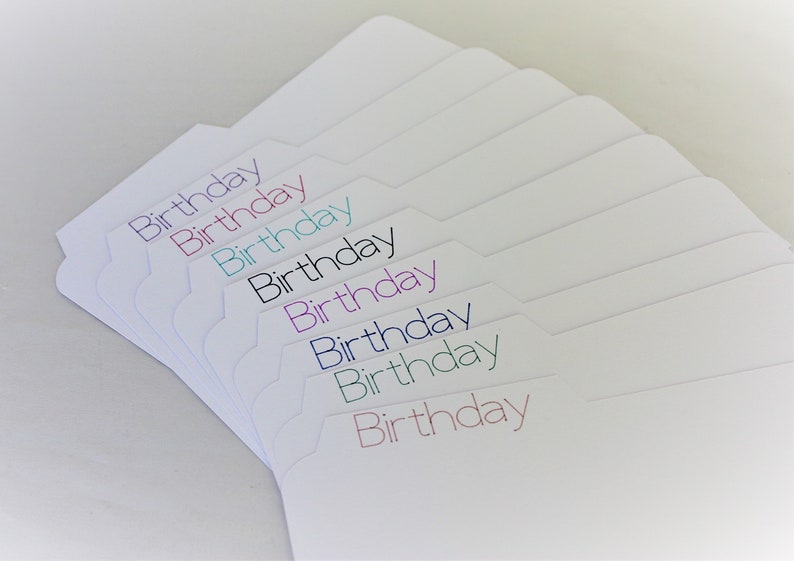 May include: A stack of white file folder tabs with the word "Birthday" printed in various colors. The tabs are arranged in a fan shape, with the text in purple, pink, teal, black, blue, green, and red.