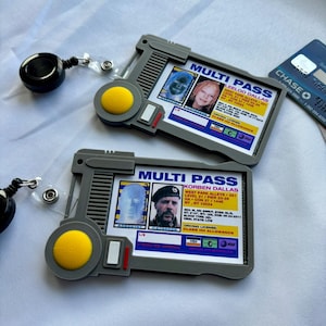 May include: Two grey plastic Multi Pass badge holders with retractable black clips. Each holder features a yellow circular button and a printed identification card. One card displays Leeloo Dallas, the other Korben Dallas, both with personal data.