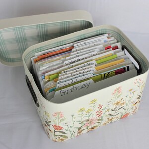 May include: A cream-colored card organizer with a floral design and a matching lid. The open box contains labeled dividers for various occasions, including "Birthday" and "Get Well Soon". The interior of the lid has a green plaid pattern.