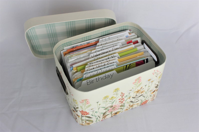 May include: A cream-colored card organizer with a floral design and a matching lid. The open box contains labeled dividers for various occasions, including "Birthday" and "Get Well Soon". The interior of the lid has a green plaid pattern.