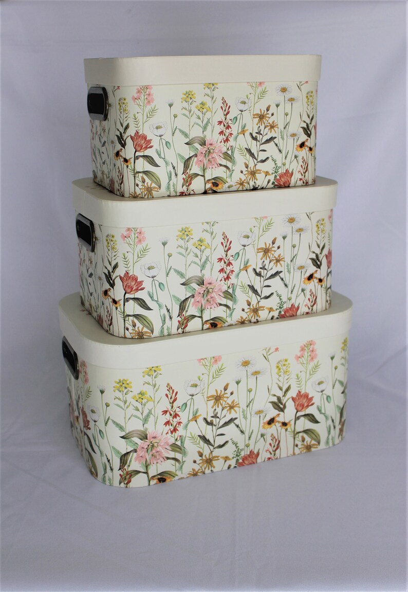 May include: Three nested storage boxes with lids, featuring a floral design of wildflowers in pink, yellow, and red on a cream background. Each box has a black handle. The boxes are oval-shaped.