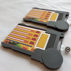 Puede incluir: Dos porta tarjetas de pl&aacute;stico gris con un dise&ntilde;o amarillo y rojo. Los porta tarjetas tienen un bot&oacute;n circular en el lateral y un lazo de pl&aacute;stico transparente para sujetar un cord&oacute;n. Los porta tarjetas tienen el texto "CLASS T" y "DNA, EAR, INS, CRE, ONE" impreso en ellos.