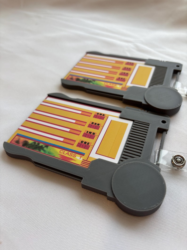 Puede incluir: Dos porta tarjetas de pl&aacute;stico gris con un dise&ntilde;o amarillo y rojo. Los porta tarjetas tienen un bot&oacute;n circular en el lateral y un lazo de pl&aacute;stico transparente para sujetar un cord&oacute;n. Los porta tarjetas tienen el texto "CLASS T" y "DNA, EAR, INS, CRE, ONE" impreso en ellos.