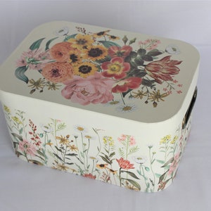 May include: Cream-colored rectangular storage box with a floral design. The lid features a bouquet of pink, orange, and yellow flowers. The sides are adorned with a wildflower pattern, and it has black handles.