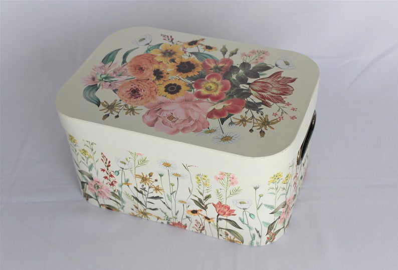 May include: Cream-colored rectangular storage box with a floral design. The lid features a bouquet of pink, orange, and yellow flowers. The sides are adorned with a wildflower pattern, and it has black handles.
