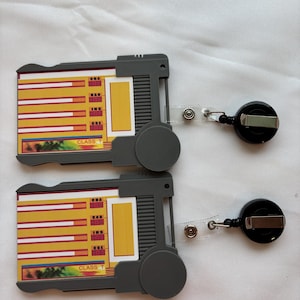 Puede incluir: Dos portagajetas de pl&aacute;stico gris con tarjetas amarillas y rojas en su interior. Las tarjetas tienen las palabras "CBA", "KAR", "INS" y "CRE" en ellas. Los portagajetas tienen cordones retr&aacute;ctiles.