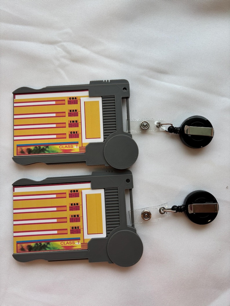 Puede incluir: Dos portagajetas de pl&aacute;stico gris con tarjetas amarillas y rojas en su interior. Las tarjetas tienen las palabras "CBA", "KAR", "INS" y "CRE" en ellas. Los portagajetas tienen cordones retr&aacute;ctiles.