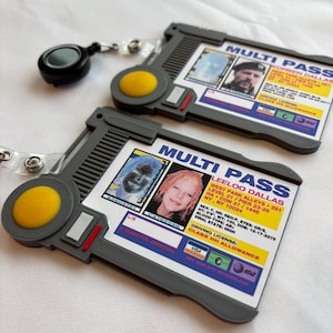 Puede incluir: Dos tarjetas multi-pass de pl&aacute;stico gris con un bot&oacute;n amarillo y un cord&oacute;n retr&aacute;ctil. Las tarjetas muestran una foto de una persona y texto que incluye "MULTI PASS", "KORBEN DALLASS", "LEELOO DALLAS", "CLASS HH ALLOWANCE" y "VISA".