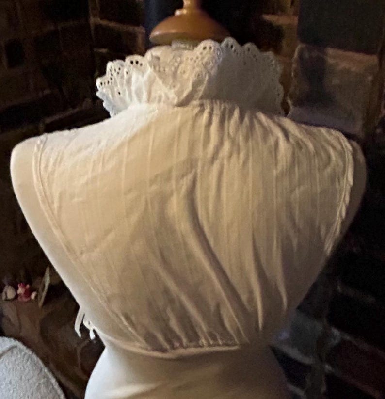 Pu&ograve; includere: Un capo bianco in stile vintage, possibilmente una camicetta o biancheria intima, esposto su un manichino. Il capo presenta un colletto a balze in pizzo traforato e pieghe verticali. Il tessuto sembra essere cotone o lino leggero.