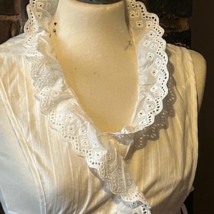 Puede incluir: Una blusa blanca sin mangas con un cuello con volantes de encaje calado. El cuello y el escote están adornados con delicados bordes festoneados. La blusa se exhibe en un maniquí, mostrando su diseño y textura.