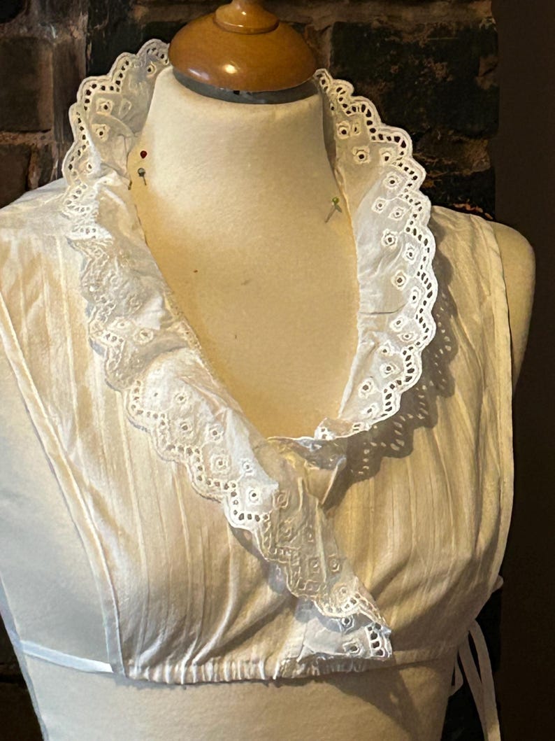 Pu&ograve; includere: Un top bianco senza maniche con un colletto a balze in pizzo traforato. Il colletto e lo scollo sono adornati da delicati bordi smerlati. Il top &egrave; esposto su un manichino, mettendo in risalto il suo design e la sua consistenza.