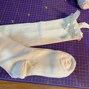 May include: Off-white knee-high socks with a ruffled trim and satin ribbon detail. The socks are made of a soft, opaque material and are laying on a purple cutting mat.