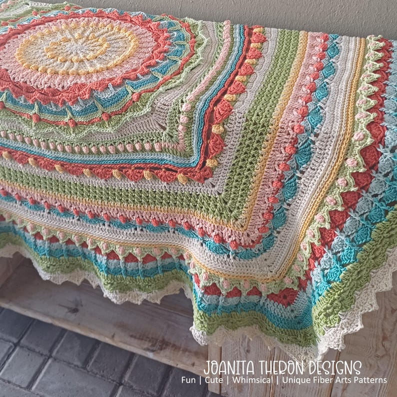 May include: A colorful, handmade crochet blanket with a circular design. The blanket features a variety of pastel colors, including pink, yellow, green, and blue. The edges are scalloped and trimmed with white. The text "JOANITA THERON DESIGNS" is visible.