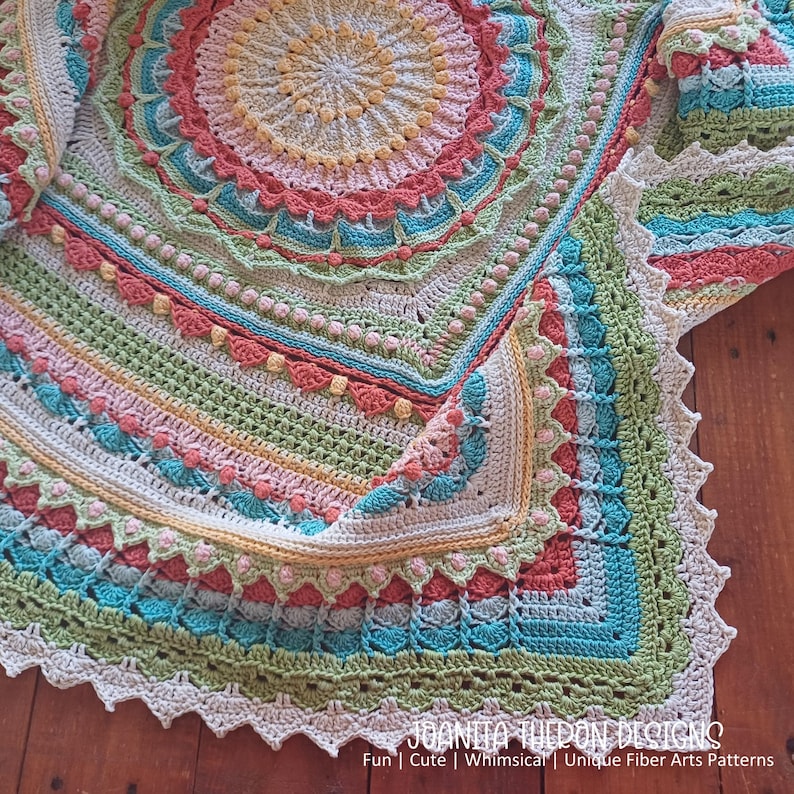 May include: A colorful, crocheted blanket with a circular design. The blanket features a variety of colors, including pink, blue, green, and yellow. The edges are scalloped, and the blanket has a textured appearance. The text "JOANITA TUEON DESIGNS" is visible at the bottom.